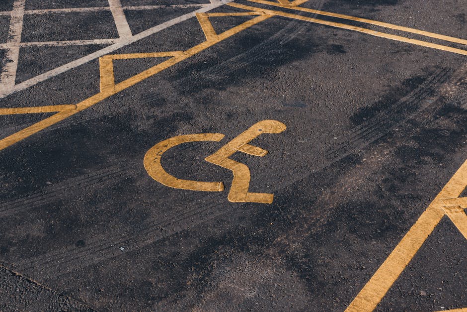 A close-up view of an asphalt parking space featuring a yellow painted accessibility symbol for disabled parking, positioned on the ground outside a residential property. Surrounding the symbol are ot